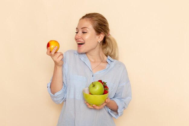 Front view young beautiful female in shirt holding plate with fruits and apple on cream wall fruit model woman pose