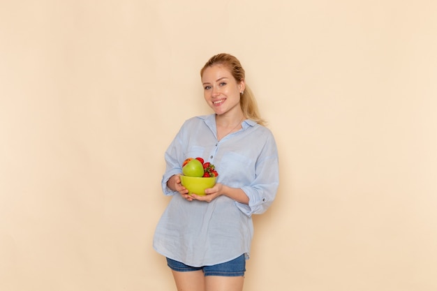 Вид спереди молодая красивая женщина в рубашке, держащая тарелку с фруктами на кремовой стене, спелая модель женщины, позирует