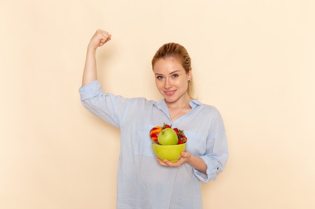 Вид спереди молодая красивая женщина в рубашке, держащая тарелку с фруктами и изгибающаяся на кремовой стене, спелая модель женщины позы
