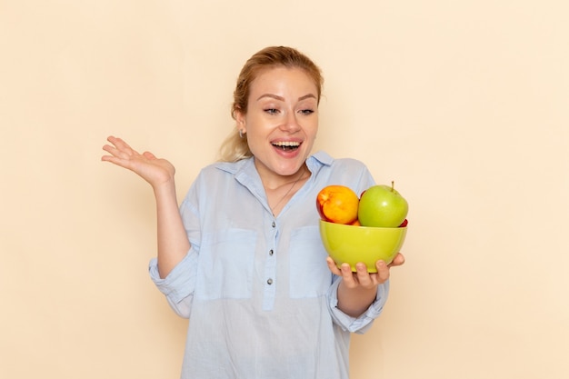 Front view young beautiful female in shirt holding plate with fruits and smiling on light-cream wall fruit model woman pose lady