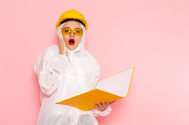 Front view young beautiful female in special white suit wearing protective helmet holding yellow files on the pink space special suit  girl woman