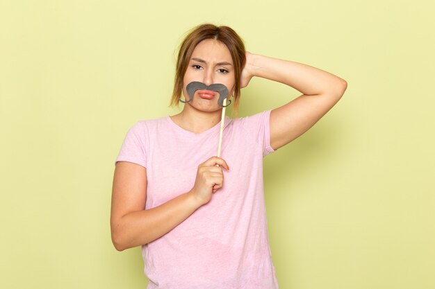 A front view young beautiful girl in pink t-shirt blue jeans posing with fake mustache and being sad on green