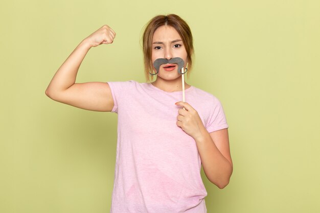 A front view young beautiful girl in pink t-shirt blue jeans posing with fake mustache and flexing on green