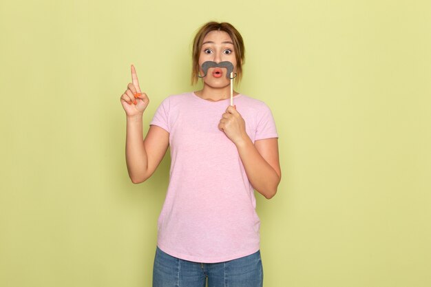 A front view young beautiful girl in pink t-shirt blue jeans posing with fake mustache on green