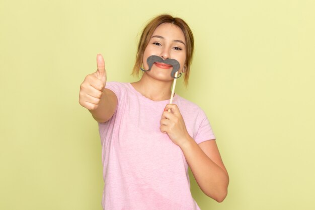 A front view young beautiful girl in pink t-shirt blue jeans posing with fake mustache and smiling on green