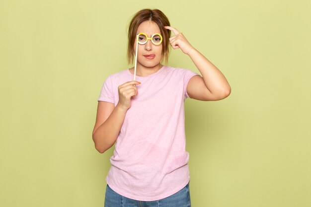 A front view young beautiful girl in pink t-shirt blue jeans posing with toy stick sunglasses on green