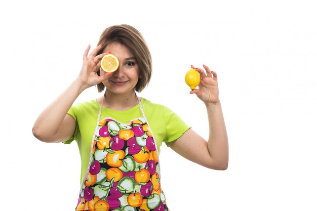 A front view young beautiful housewife in green shirt colorful cape holding covering her eye with sliced lemon on the white background house female kitchen