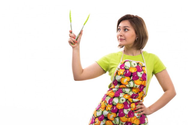 A front view young beautiful housewife in green shirt colorful cape holding green kitchen appliance smiling on the white background house cleaning kitchen