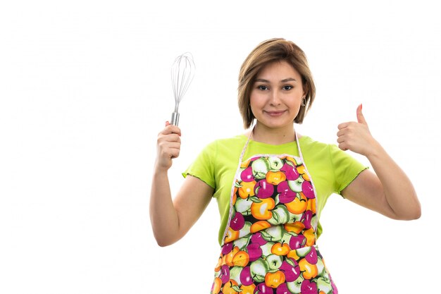 A front view young beautiful housewife in green shirt colorful cape holding kitchen appliance smiling on the white background house cleaning kitchen