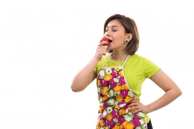 A front view young beautiful housewife in green shirt colorful cape holding tasting red tomatoes smiling on the white background house cleaning kitchen