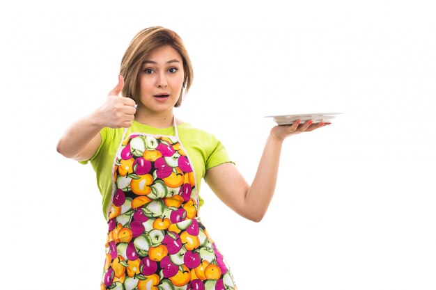 A front view young beautiful housewife in green shirt colorful cape holding white plate smiling on the white background house cleaning kitchen