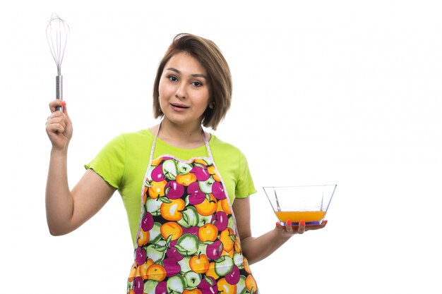 白い背景の家の女性のキッチンで食事を準備する液体の卵を混合緑のシャツカラフルなケープの正面の若い美しい主婦