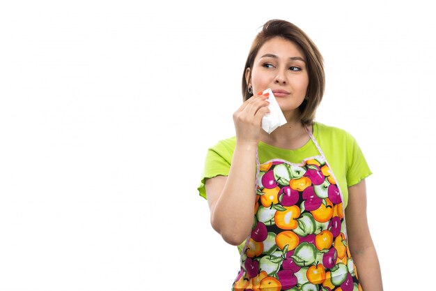 A front view young beautiful housewife in green shirt colorful cape posing cleanign her mouth smiling happy on the white background house female kitchen