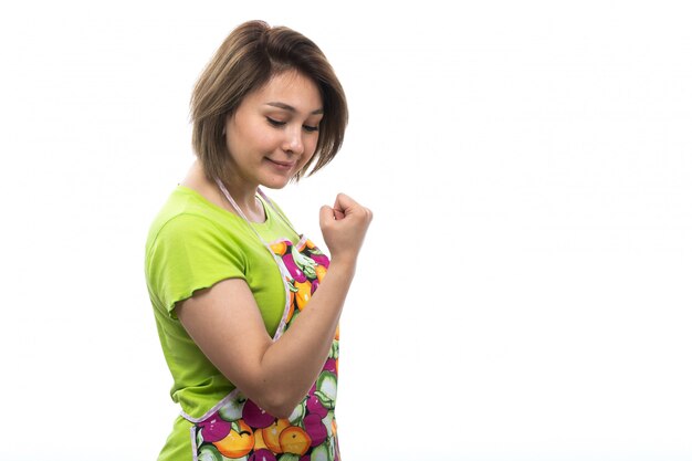 A front view young beautiful housewife in green shirt colorful cape posing happy smiling on the white background house female kitchen