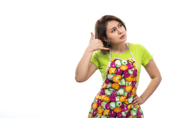 A front view young beautiful housewife in green shirt colorful cape posing showing imaginary phone talk on the white background house female kitchen
