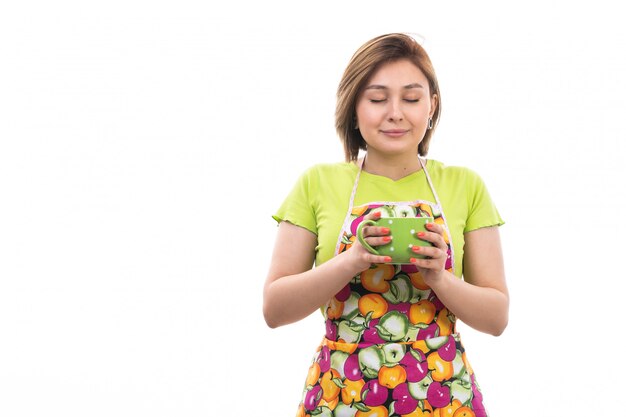 A front view young beautiful housewife in green shirt colorful cape smiling holding green cup on the white background house cleaning kitchen