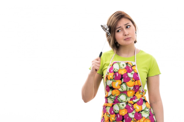 Вид спереди молодая красивая домохозяйка в зеленой рубашке красочный плащ улыбается, держа кухонный прибор, думая на белом фоне уборка дома кухня
