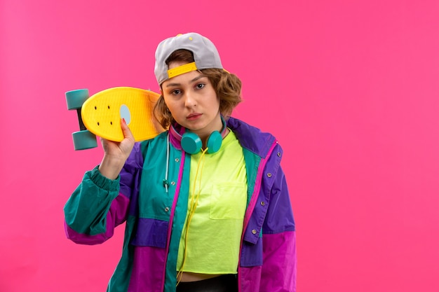 A front view young beautiful lady in acid colored shirt black trousers colorful jacket with blue earphones holding skateboard