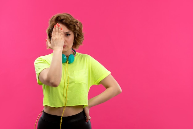 A front view young beautiful lady in acid colored shirt black trousers with blue earphones covering her eye