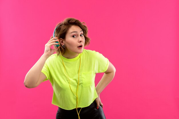 A front view young beautiful lady in acid colored shirt black trousers with blue earphones listening to music trying to hear