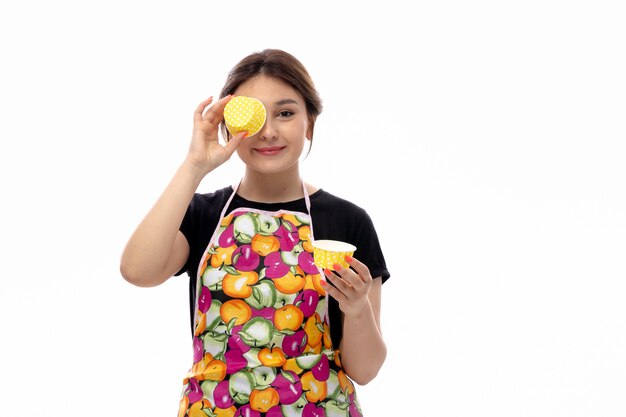 A front view young beautiful lady in black shirt and colorful cape holding yellow little cake pans smiling covering her eye