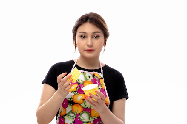 A front view young beautiful lady in black shirt and colorful cape holding yellow little cake pans