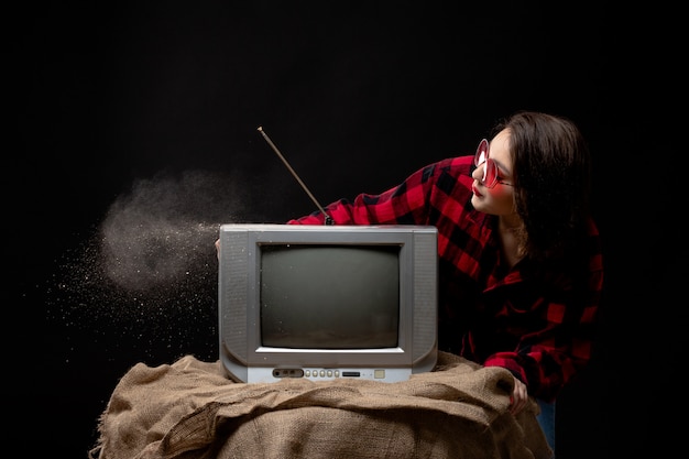 Free photo a front view young beautiful lady in checkered red-black shirt in red sunglasses near little tv blowing out dust