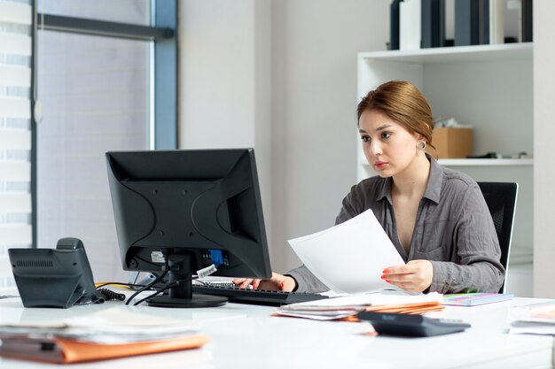 Вид спереди молодая красивая дама в серой рубашке работает на своем компьютере, сидя в своем офисе во время дневного строительства