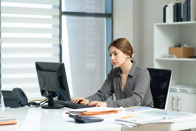 Una giovane bella signora di vista frontale in camicia grigia che lavora al suo pc che si siede dentro il suo ufficio durante l'attività di lavoro di costruzione di giorno