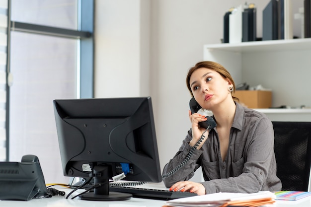 A front view young beautiful lady in grey shirt working on her pc sitting inside her office talking on the phone during daytime building job activity