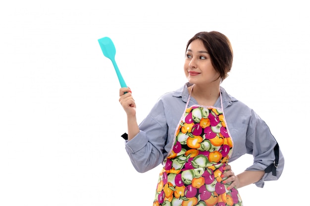 Вид спереди молодая красивая дама в светло-голубой рубашке и красочной накидке, думая, что держит синий кухонный прибор улыбается