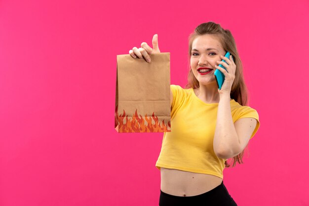 A front view young beautiful lady in orange colored shirt black trousers smiling talking on the phone holding package