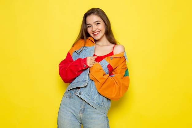 A front view young beautiful lady in red shirt colorful coat and blue jeans posing and smiling
