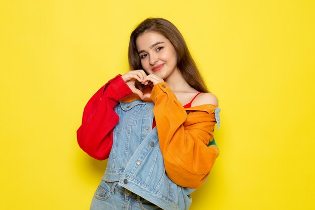 A front view young beautiful lady in red shirt colorful coat and blue jeans showing heart sign model girl color female