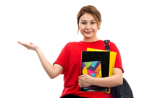 A front view young beautiful lady in red t-shirt black jeans holding different copybooks and files smiling with bag on the white