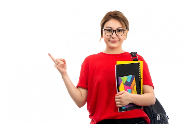 A front view young beautiful lady in red t-shirt black jeans holding different copybooks and files smiling with bag on the white