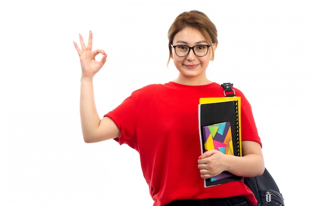 A front view young beautiful lady in red t-shirt black jeans holding different copybooks and files smiling with bag on the white
