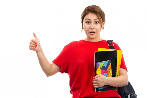Вид спереди молодая красивая дама в красной футболке и черных джинсах держит различные тетради и файлы с сумкой на белом