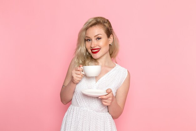 彼女の顔に笑顔でお茶のカップを保持している白いドレスの正面の若い美しい女性