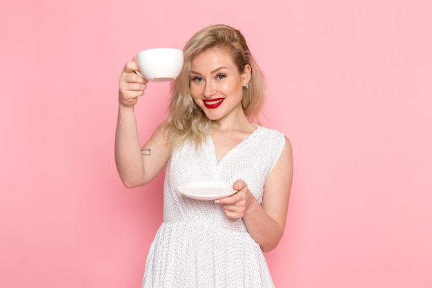 A front view young beautiful lady in white dress holding cup of tea with smile on her face