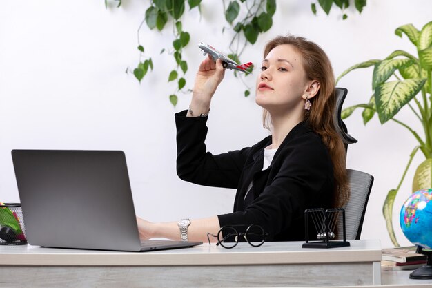 A front view young beautiful lady in white shirt and black jacket using her laptop holding toy plane in front of table with leaves hanging