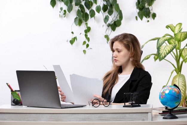 A front view young beautiful lady in white shirt and black jacket working with documents using her laptop in front of table with leaves hanging