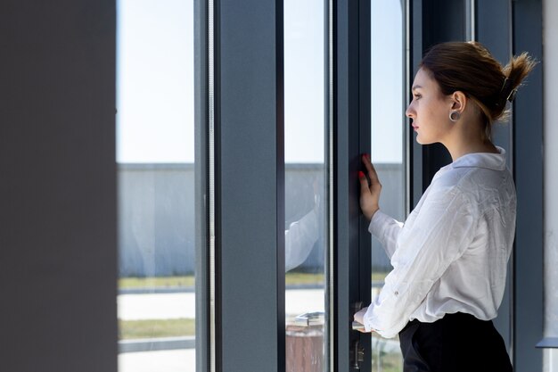 A front view young beautiful lady in white shirt black trousers looking at the distance through window in the hall waiting during daytime building job activity