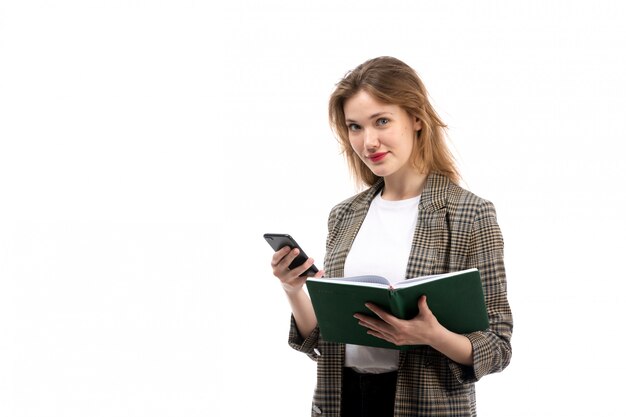 A front view young beautiful lady in white t-shirt black jeans and coat holding black smartphone and green book on the white