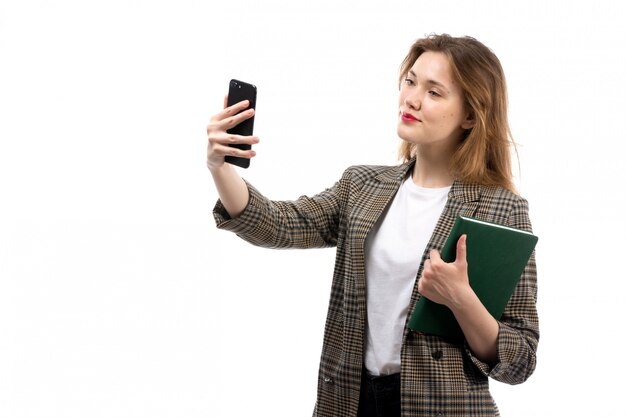 A front view young beautiful lady in white t-shirt black jeans and coat holding black smartphone taking a selfie and green book on the white