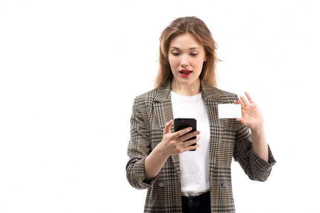 A front view young beautiful lady in white t-shirt black jeans and coat holding black smartphone and white card on the white