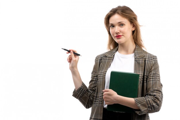 A front view young beautiful lady in white t-shirt black jeans and coat holding green book and pen smiling on the white