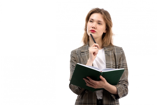 A front view young beautiful lady in white t-shirt black jeans and coat holding green book writing down thinking on the white