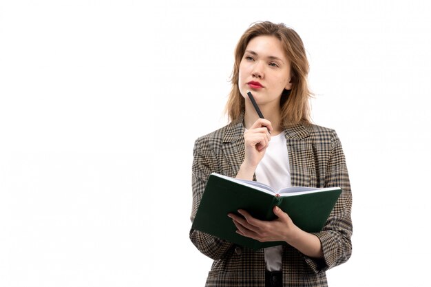 A front view young beautiful lady in white t-shirt black jeans and coat holding green book writing down thinking on the white