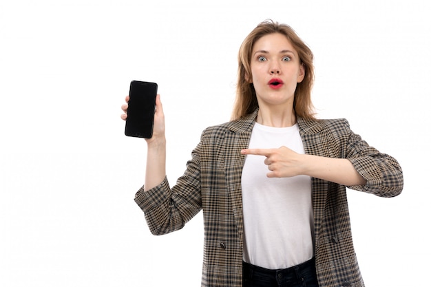 A front view young beautiful lady in white t-shirt black jeans and coat holding showing smartphone surprised on the white